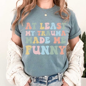 May include: A blue t-shirt with the text "At Least My Trauma Made Me Funny" in rainbow colours.
