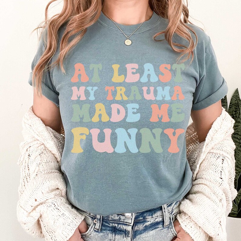 May include: A blue t-shirt with the text "At Least My Trauma Made Me Funny" in rainbow colours.