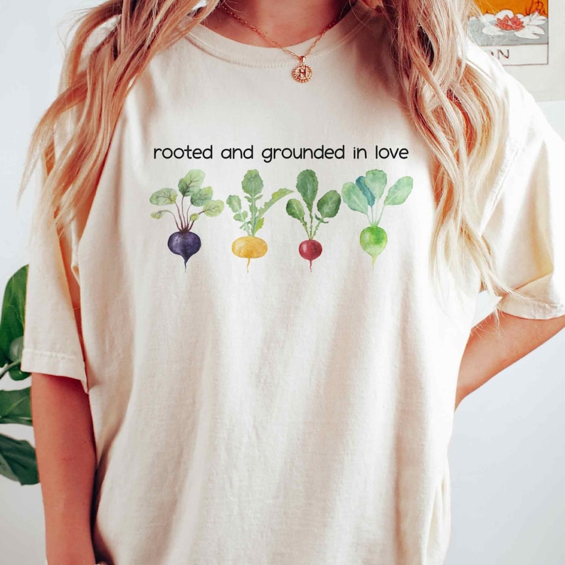 May include: A white t-shirt with the text "rooted and grounded in love" printed on the front. The text is surrounded by a row of four watercolor illustrations of different colored root vegetables.