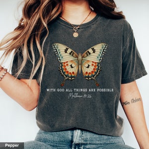 Puede incluir: Una camiseta gris oscuro con un gr&aacute;fico de mariposa de estilo vintage y el texto "With God all things are possible Matthew 19:26".