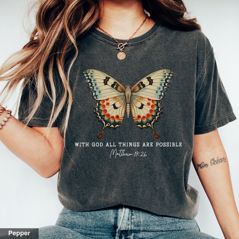 Puede incluir: Una camiseta gris oscuro con un gr&aacute;fico de mariposa de estilo vintage y el texto "With God all things are possible Matthew 19:26".