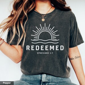 Può includere: Una maglietta grigia scura con una grafica bianca di un sole che sorge sopra l'acqua. Il testo "REDEEMED" è sotto la grafica, con il riferimento biblico "EPHESIANS 1:7" sotto.