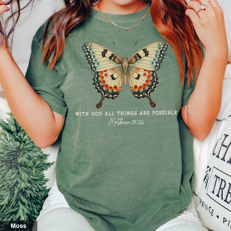 Puede incluir: Una camiseta verde musgo con un gr&aacute;fico de mariposa colorida y el texto "Con Dios todo es posible Mateo 19:26".