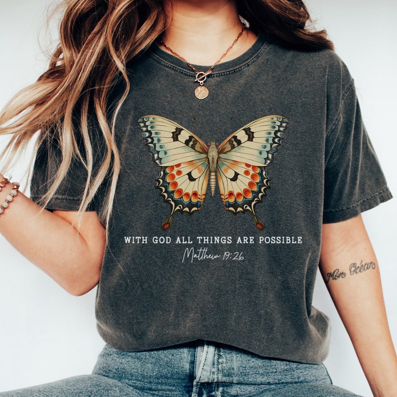 Puede incluir: Una camiseta gris oscuro con un gr&aacute;fico de mariposa colorido y el texto "Con Dios todo es posible Mateo 19:26".