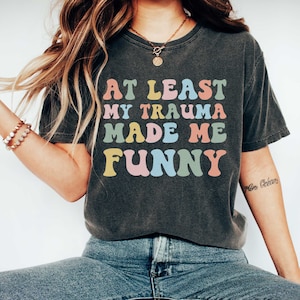 May include: A dark grey t-shirt with the text "At Least My Trauma Made Me Funny" in colourful, retro-style lettering.