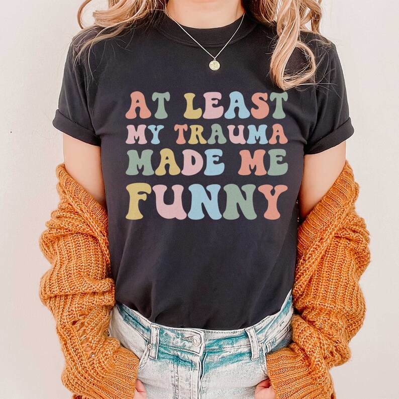 May include: A black t-shirt with the text "At Least My Trauma Made Me Funny" in rainbow colours.