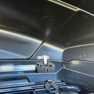 May include: Close-up of a black roof box with a white plastic latch and a black strap. The roof box is open and the inside is visible.
