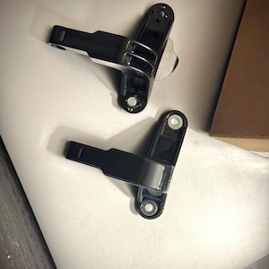 May include: Two black metal hinges with silver accents. The hinges are in a closed position.