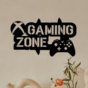 May include: A black metal sign with the words "Gaming Zone" and a stylized image of a video game controller. A laptop computer with a blank screen is on a white table in front of the sign.