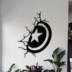 Op de afbeelding: Zwarte metalen wanddecoratie met een gestileerd ster- en cirkelontwerp, dat lijkt op een superheldenschild, met een gebarsten muureffect. De decoratie is bevestigd aan een witte muur met potplanten op de voorgrond.