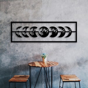 Pode incluir: Decoração de parede de metal preto com uma série de luas crescentes em diferentes fases. A decoração está montada em uma parede de concreto cinza acima de uma mesa de madeira e dois banquinhos de madeira.