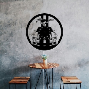 May include: Black metal wall clock with a car and a horse design. The clock has Roman numeral markings and a black background. The clock is hanging on a gray wall with two wooden tables in front of it.