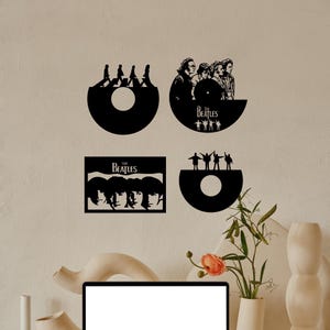 May include: Four black vinyl record wall art pieces featuring The Beatles. The first piece shows the band members walking across a crosswalk. The second piece shows the band members in a portrait. The third piece shows the band members in a portrait with the text "The Beatles" below. The fourth piece shows the band members in a portrait with the text "The Beatles" above.