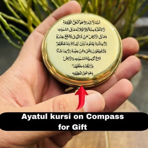 May include: A gold compass with Arabic text inscribed on the face. The text is the Ayatul Kursi, a verse from the Quran. The compass is being held in a hand.