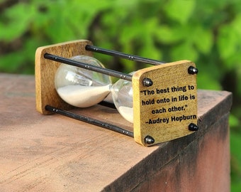 Op de afbeelding: Zandloper met een houten frame en heldere glazen bollen gevuld met wit zand. Het houten frame heeft de quote "Het beste in het leven is elkaar vasthouden." -Audrey Hepburn.