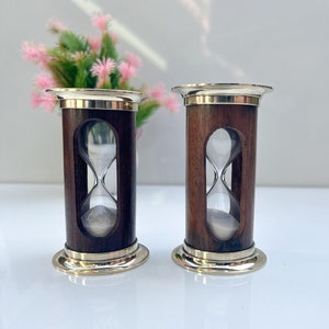 以下が含まれることがあります： 真鍮のアクセントが付いた木製の砂時計タイマー2つ。砂は白く、ガラスは透明です。砂時計は白い表面の上にあります。