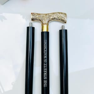 Puede incluir: Un bastón negro con un mango dorado adornado. El mango tiene forma de T y está grabado con un diseño floral. El bastón está grabado con el texto "THE STREETZ IS WATCHING".