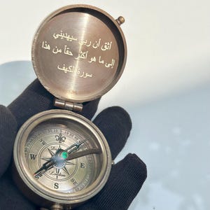 Könnte beinhalten: Ein antiker Messingkompass mit offenem Deckel, der arabische Schrift zeigt. Das Kompasszifferblatt zeigt Richtungsmarkierungen und das Wort "London". Der Kompass wird vor einem dunklen Hintergrund gehalten.