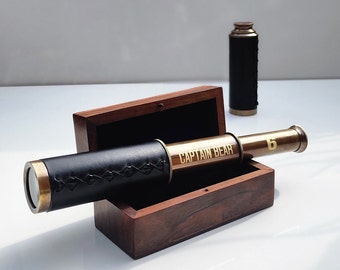 May include: A brass telescope with a black leather covering and the words "Captain Bear" engraved on it. The telescope is in a wooden box with a hinged lid.