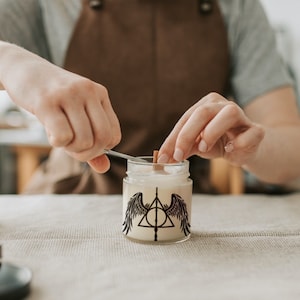 Puede incluir: Una persona est&aacute; a&ntilde;adiendo una mecha de madera a un tarro de vela. El tarro de vela tiene un dise&ntilde;o en blanco y negro de un tri&aacute;ngulo con un c&iacute;rculo en su interior y alas a cada lado. El dise&ntilde;o recuerda al s&iacute;mbolo de las Reliquias de la Muerte de la serie Harry Potter.