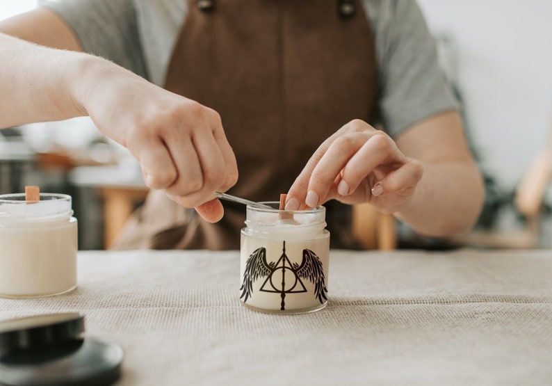 Puede incluir: Una persona est&aacute; a&ntilde;adiendo una mecha de madera a un tarro de vela. El tarro de vela tiene un dise&ntilde;o en blanco y negro de un tri&aacute;ngulo con un c&iacute;rculo en su interior y alas a cada lado. El dise&ntilde;o recuerda al s&iacute;mbolo de las Reliquias de la Muerte de la serie Harry Potter.
