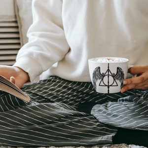 Puede incluir: Una persona con pantalones a rayas y una sudadera blanca est&aacute; sentada en una alfombra blanca, sosteniendo una taza blanca con un s&iacute;mbolo negro de las Reliquias de la Muerte y alas. La taza est&aacute; llena de una bebida caliente y malvaviscos. La persona tambi&eacute;n est&aacute; sosteniendo un libro.