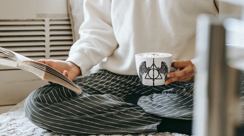 Puede incluir: Una persona con pantalones a rayas y una sudadera blanca est&aacute; sentada en una alfombra blanca, sosteniendo una taza blanca con un s&iacute;mbolo negro de las Reliquias de la Muerte y alas. La taza est&aacute; llena de una bebida caliente y malvaviscos. La persona tambi&eacute;n est&aacute; sosteniendo un libro.