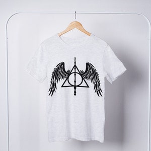Puede incluir: Una camiseta blanca con un gr&aacute;fico negro del s&iacute;mbolo de las Reliquias de la Muerte con alas. El s&iacute;mbolo es un tri&aacute;ngulo con un c&iacute;rculo dentro y una varita que atraviesa el c&iacute;rculo.