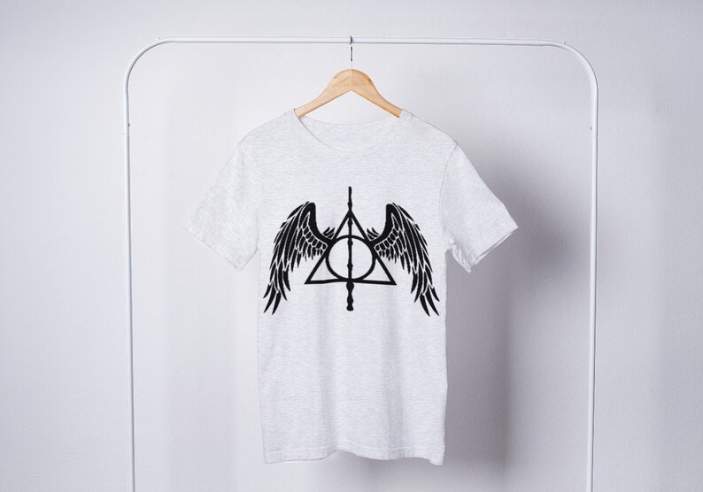 Puede incluir: Una camiseta blanca con un gr&aacute;fico negro del s&iacute;mbolo de las Reliquias de la Muerte con alas. El s&iacute;mbolo es un tri&aacute;ngulo con un c&iacute;rculo dentro y una varita que atraviesa el c&iacute;rculo.