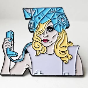 Può includere: Spilla smaltata raffigurante una donna stilizzata con capelli biondi e rossetto rosso, che indossa un copricapo blu e tiene in mano un telefono blu. La spilla ha uno sfondo bianco e lilla chiaro.