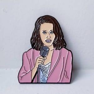 May include: A pink enamel pin featuring a woman with brown hair holding a microphone. She is wearing a pink jacket and a white lace top.