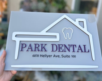 May include: A white sign with a house outline and a tooth inside. The text "PARK DENTAL" is in purple lettering with the address "6070 Hellyer Ave, Suite 100" below.