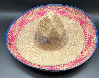 May include: A straw sombrero with a woven design. The hat features a blue brim and a pink and red patterned band. The crown is adorned with blue decorative stitching. A traditional Mexican hat.