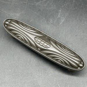 May include: A dark brown, decorative, metal cabinet pull with an intricate, wave-like design.