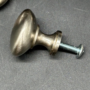 May include: A silver-toned cabinet knob with a round, slightly oval-shaped top and a screw. The knob has a brushed finish.