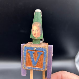 May include: A wooden toy robot with a green hat featuring a portrait of a person. The robot has a blue letter "V" on its chest and purple arms.