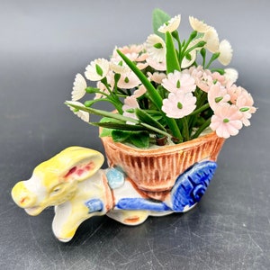 May include: A ceramic donkey figurine pulling a wagon with a planter filled with pink and white flowers. The donkey is painted yellow, blue, and red. The wagon is painted brown, blue, and white.