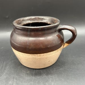 May include: A handmade ceramic jar with a handle. The jar is two-toned, with a dark brown upper section and a tan lower section. The jar has a round body and a wide opening, perfect for storing items.