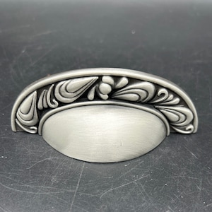 May include: A silver cabinet pull with a decorative floral design. The pull is shaped like a half-circle with a rounded oval in the center.