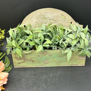 May include: A rustic green metal planter with a distressed finish. The planter has a curved top and is filled with artificial green plants.