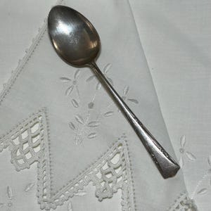 May include: A silver-toned metal spoon featuring a polished oval bowl and a slender handle decorated with an embossed floral pattern along the edges. The spoon has a reflective surface and a tapered end, showcasing a classic design with intricate detailing.