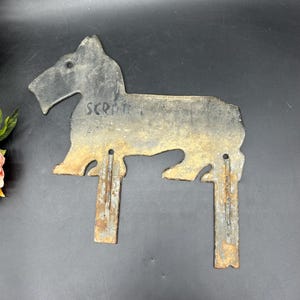May include: A metal silhouette of a Scottish Terrier dog with two metal stakes for mounting. The silhouette is painted black and has a weathered, rusty appearance. The text "SCRPT." is visible on the dog's body.