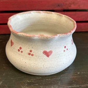 May include: A small, handmade ceramic bowl with a speckled, off-white glaze. The rim is painted with a reddish-pink color, and the bowl is decorated with red hearts and dots. The bowl has a slightly irregular shape, giving it a rustic, handcrafted feel.