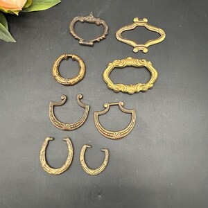 May include: A collection of antique brass drawer pulls in various shapes and designs. The pulls include ornate oval, circular, and horseshoe-shaped pieces, all with a vintage aesthetic. The hardware is arranged on a dark surface, showcasing their intricate details.