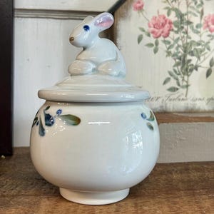 May include: A white ceramic jar with a lid, featuring a bunny figurine on top. The jar is decorated with blue and green floral accents. The bunny has pink ears and blue eyes. The jar is sitting on a wooden surface.