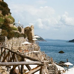 May include: A scenic coastal view featuring a rocky cliffside with stone steps and wooden railings leading down to the water. White umbrellas provide shade. The blue sea stretches to the horizon, with an island in the distance.