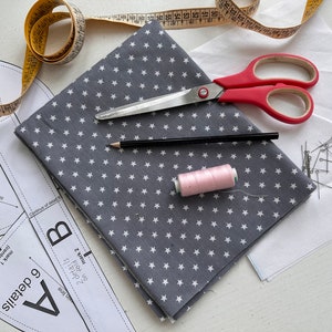 K&ouml;nnte beinhalten: Eine flache Anordnung von N&auml;hzubeh&ouml;r, darunter ein grauer Stoff mit wei&szlig;en Sternen, eine rosa Garnrolle, ein Bleistift, eine Schere, ein Ma&szlig;band und N&auml;hnadeln auf einer wei&szlig;en Oberfl&auml;che. Der Stoff ist in der H&auml;lfte gefaltet und der Text "6 details A" und "2 details B" ist auf dem Stoff sichtbar.