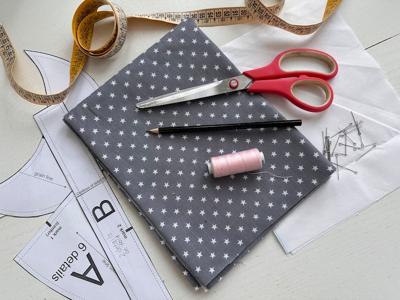 K&ouml;nnte beinhalten: Eine flache Anordnung von N&auml;hzubeh&ouml;r, darunter ein grauer Stoff mit wei&szlig;en Sternen, eine rosa Garnrolle, ein Bleistift, eine Schere, ein Ma&szlig;band und N&auml;hnadeln auf einer wei&szlig;en Oberfl&auml;che. Der Stoff ist in der H&auml;lfte gefaltet und der Text "6 details A" und "2 details B" ist auf dem Stoff sichtbar.