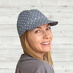 K&ouml;nnte beinhalten: Eine graue und wei&szlig;e Baseballkappe mit Sternenmuster und einem gebogenen Schirm. Die Kappe wird von einer Person mit blonden Haaren getragen.