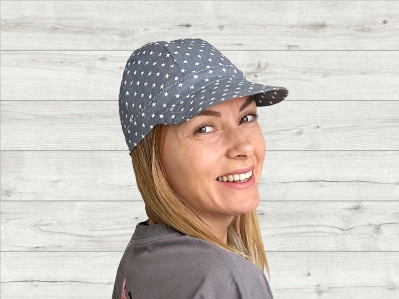 K&ouml;nnte beinhalten: Eine graue und wei&szlig;e Baseballkappe mit Sternenmuster und einem gebogenen Schirm. Die Kappe wird von einer Person mit blonden Haaren getragen.
